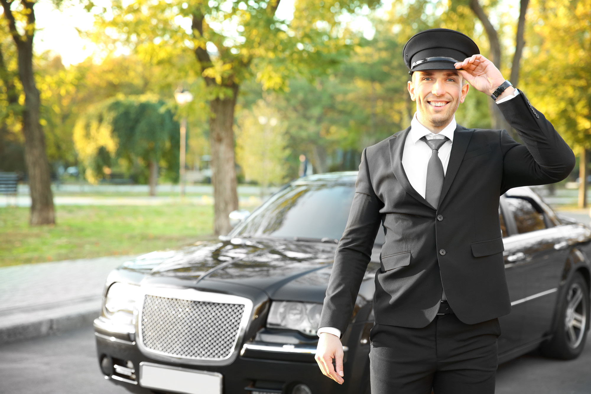 Young chauffeur adjusting hat near luxury car on the street